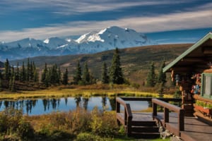 Wooden lodge near pond with snow covered mountain in background.