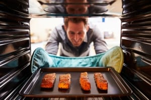 A smiling man eagerly pulls out his baked salmon from the oven while wearing heat protection on his hands.